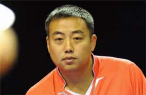 Sportler mit grauem Haar fokussiert im Wettkampf, trägt oranges Shirt, Bewegung angedeutet.