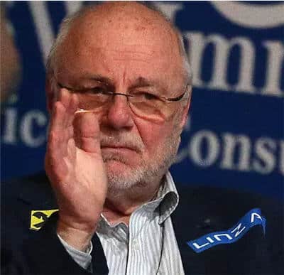 Älterer Mann mit Geste bei Konferenz, nachdenklich, formell gekleidet, verschwommener Hintergrund.