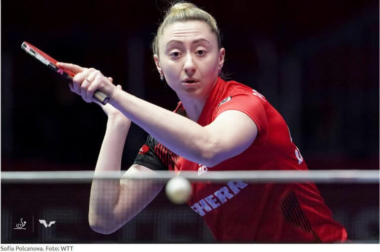Tischtennisspielerin fokussiert bei Turnier, bereit zum Rückhand-Schlag im roten Trikot.