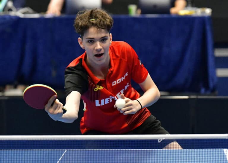 Junger Tischtennisspieler fokussiert beim Aufschlag auf internationalem Turnier, rot-schwarzes Trikot, dynamisch.