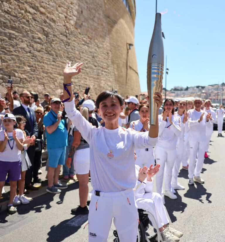 Frau mit Fackel beim olympischen Lauf, feiernd in sonniger Kulisse vor alter Steinmauer.