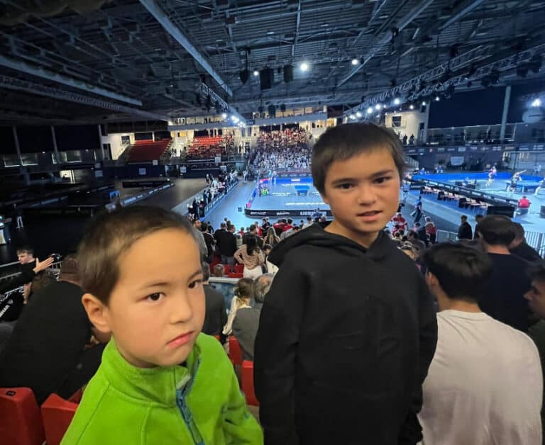 Tischtennisturnier in großer Halle mit zwei Jungen im Vordergrund, lebhaftes Publikum in roten Sitzen.
